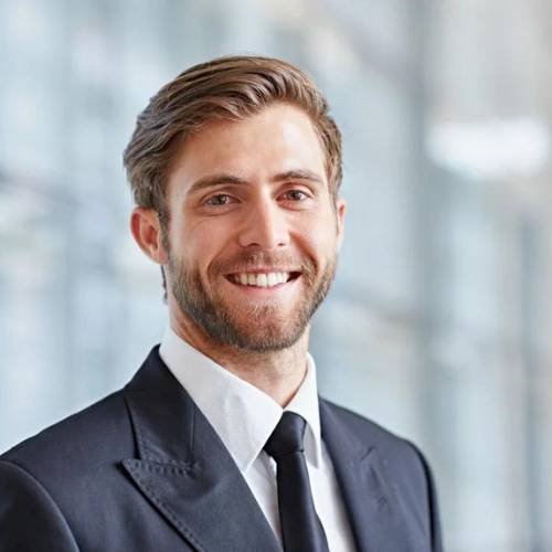 A man with short brown hair and a beard is smiling at the camera. He is wearing a black suit, a white shirt, and a black tie. The background is blurred, featuring a bright interior with large windows.